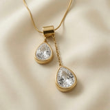 Duo Larme Crystal Asymmetric Pendant Necklace - macro close-up on ivory silk showing two bezel-set pear-cut crystal zirconia at asymmetric heights, brilliant white facets and gold bezels in full detail