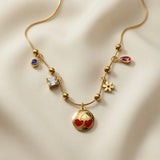 Cerise Charm Station Necklace - close-up flat lay on ivory silk showing red enamel cherry medallion centrepiece, gold daisy, ruby-rose pear crystal drop and princess white crystal on gold ball station chain