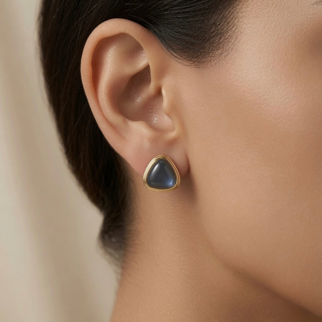Orage Bleu Triangle Earrings - worn on model ear showing rounded-triangle slate blue-grey cabochon stud in ridged gold bezel filling lobe with quiet luxury