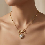 Noeud Coeur Crystal Bow Necklace - worn on mannequin collarbone showing gold bow at chest centre, swinging crystal heart halo pendant and satellite ball chain against skin
