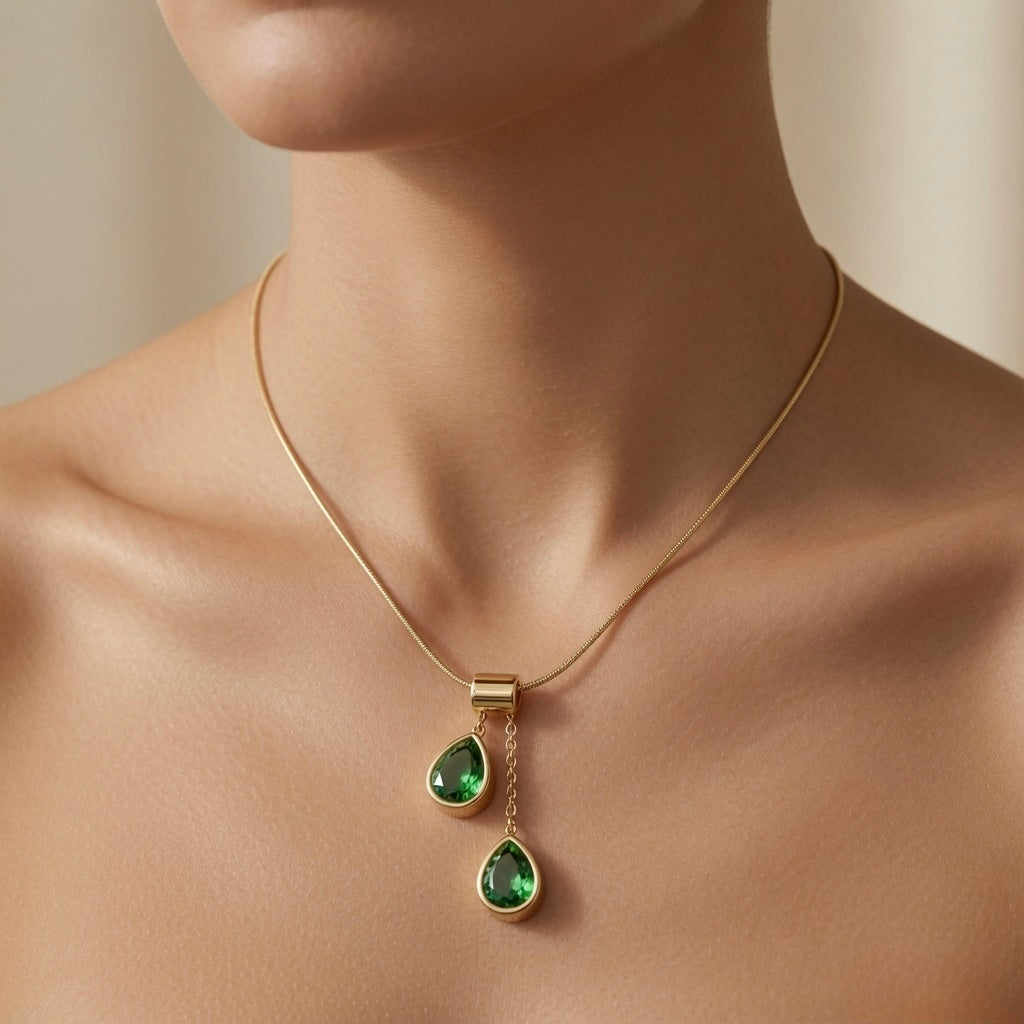 Duo Larme Émeraude Asymmetric Pendant Necklace - worn on mannequin collarbone showing upper emerald pear at collarbone and lower emerald pear at sternum, two green teardrops glowing against warm skin