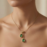 Duo Larme Émeraude Asymmetric Pendant Necklace - worn on mannequin collarbone showing upper emerald pear at collarbone and lower emerald pear at sternum, two green teardrops glowing against warm skin