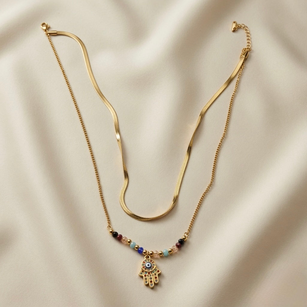 Hamsa Gemstone Layer Necklace Set - full flat lay on ivory silk showing two necklaces: flat herringbone snake chain above and multicolour gemstone bead station chain with large multicolour pavé Hamsa hand pendant below, both with lobster clasps and curb extenders