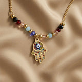 Hamsa Gemstone Layer Necklace Set - macro close-up on taupe silk showing large multicolour pavé Hamsa hand pendant with red, teal, green, champagne and white crystal mosaic and navy blue enamel evil eye at palm centre, with multicolour faceted gemstone beads and gold ball stations on curb chain