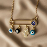 Nazar Pin Charm Necklace - macro close-up on taupe silk showing large gold safety pin with navy blue evil eye disc in pavé crystal zirconia halo at clasp end, white enamel evil eye bead, black enamel evil eye bead, pavé crystal Hamsa hand with evil eye at palm and turquoise enamel evil eye bead sliding on pin bar
