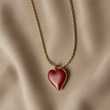 Grand Coeur Rouge Enamel Heart Pendant Necklace - close-up flat lay on taupe silk showing large burgundy-red glossy enamel puffy heart pendant in gold bezel on box chain, high-gloss lacquer surface and warm gold frame detail visible