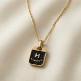 Initial Black Enamel Tag Pendant Necklace - macro close-up on ivory silk showing rounded-corner rectangular black enamel pendant in warm gold frame with bold raised gold letter H at centre and capital letter stamp below, on delicate gold cable chain with smooth gold bail