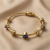 Nazar Cable Wire Evil Eye Station Bangle - flat lay on champagne silk showing complete twisted gold cable wire bangle with three deep blue evil eye disc stations in cylindrical gold barrel bezels alternating with three crystal pavé rondelle stations flanked by gold disc collars, gold barrel magnetic clasp at top