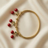 Cerise Charm Drop Tube Bangle - full flat lay on ivory silk showing complete smooth gold tube bangle with three cherry charm drops hanging from gold ring stations on front arc: each charm a pair of deep burgundy-red round beads on gold Y-fork stem with four-leaf green enamel leaf cluster at top, hinge clasp visible at back