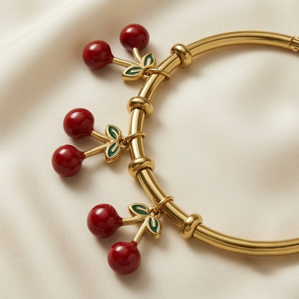 Cerise Charm Drop Tube Bangle - macro close-up on ivory silk showing three cherry charm drops in detail: paired deep burgundy-red round beads on gold Y-fork stems with four-leaf deep forest green enamel leaf clusters with lighter green vein detail, hanging from gold ring stations on smooth mirror-polished gold tube bangle