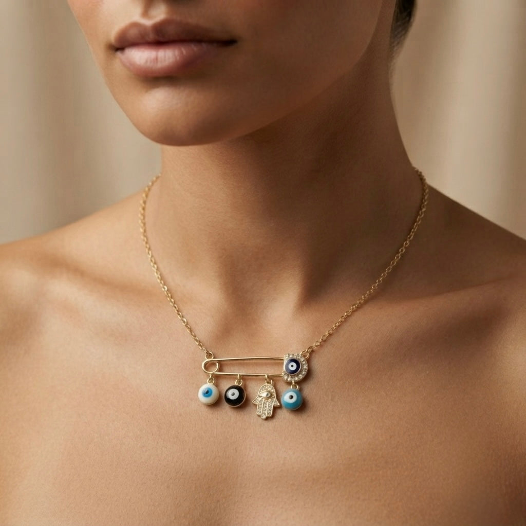 Nazar Pin Charm Necklace - worn on model bare collarbone showing gold safety pin centrepiece with navy pavé evil eye at clasp, four sliding protective charms: white evil eye bead, black evil eye bead, pavé Hamsa hand and turquoise evil eye bead hanging below pin bar on delicate gold cable chain against warm skin
