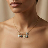 Nazar Pin Charm Necklace - worn on model bare collarbone showing gold safety pin centrepiece with navy pavé evil eye at clasp, four sliding protective charms: white evil eye bead, black evil eye bead, pavé Hamsa hand and turquoise evil eye bead hanging below pin bar on delicate gold cable chain against warm skin