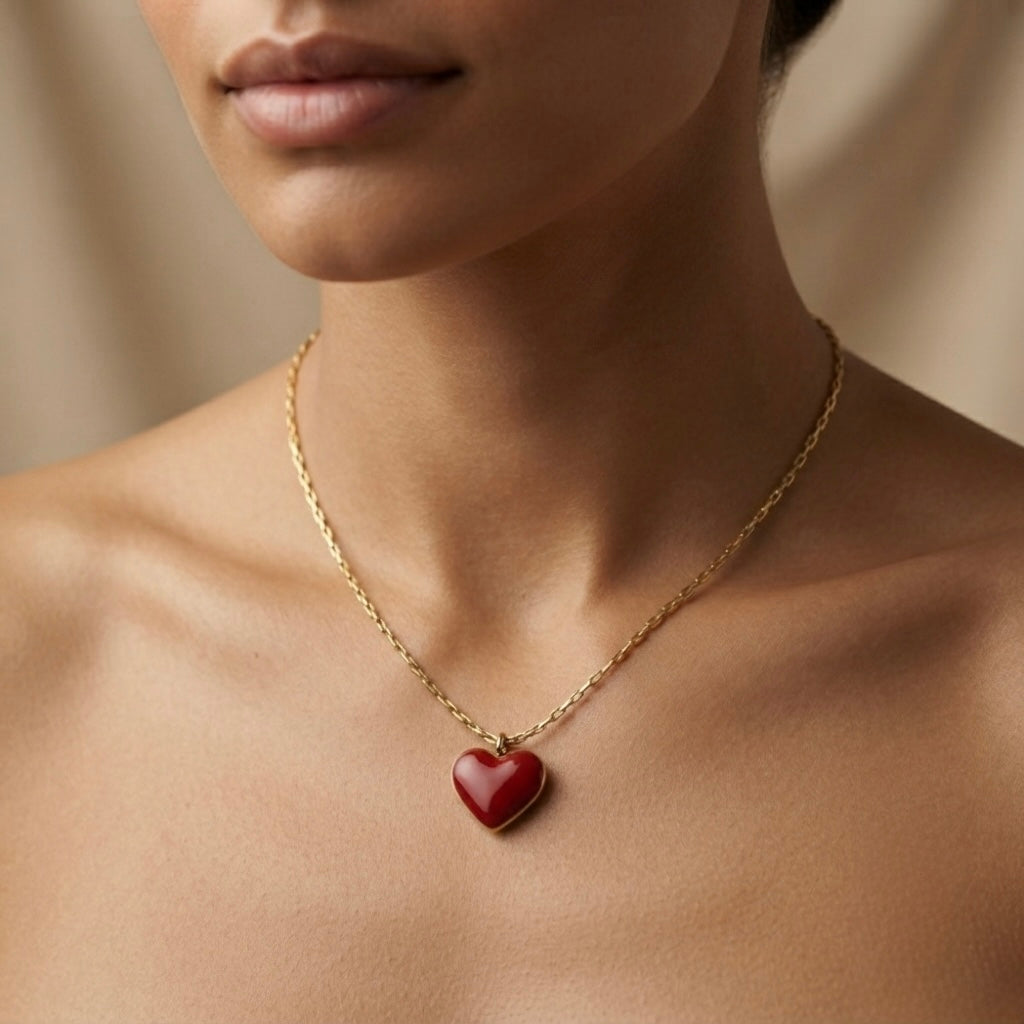 Grand Coeur Rouge Enamel Heart Pendant Necklace - worn on model sternum against warm dark skin showing large burgundy-red glossy enamel puffy heart pendant in gold bezel resting at chest centre on delicate gold box chain, high-gloss lacquer surface catching light