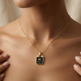 Initial Black Enamel Tag Pendant Necklace - worn on model sternum with both hands framing pendant, showing black enamel tag with raised gold H initial resting at chest centre on delicate gold cable chain against warm skin