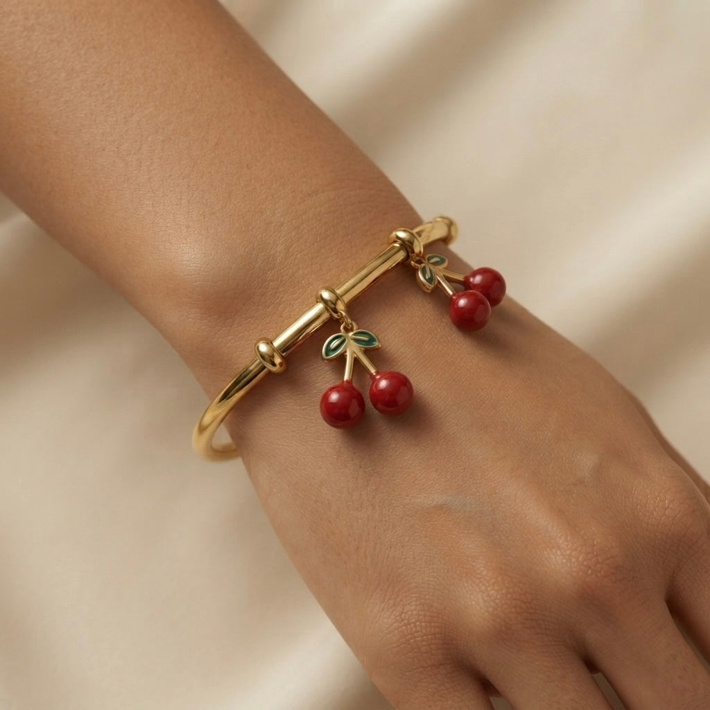 Cerise Charm Drop Tube Bangle - worn on model wrist showing two cherry charm drops hanging freely below smooth gold tube bangle: paired deep red round beads on gold Y-fork stems with green enamel leaf clusters swinging against warm skin on ivory silk background