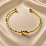 Nœud d’Amour Love Knot Ball-End Cuff Bangle - overhead view on champagne silk showing complete open cuff bangle from above: smooth round-wire gold arc with sculptural love knot at base centre and two smooth polished gold ball finials framing open back