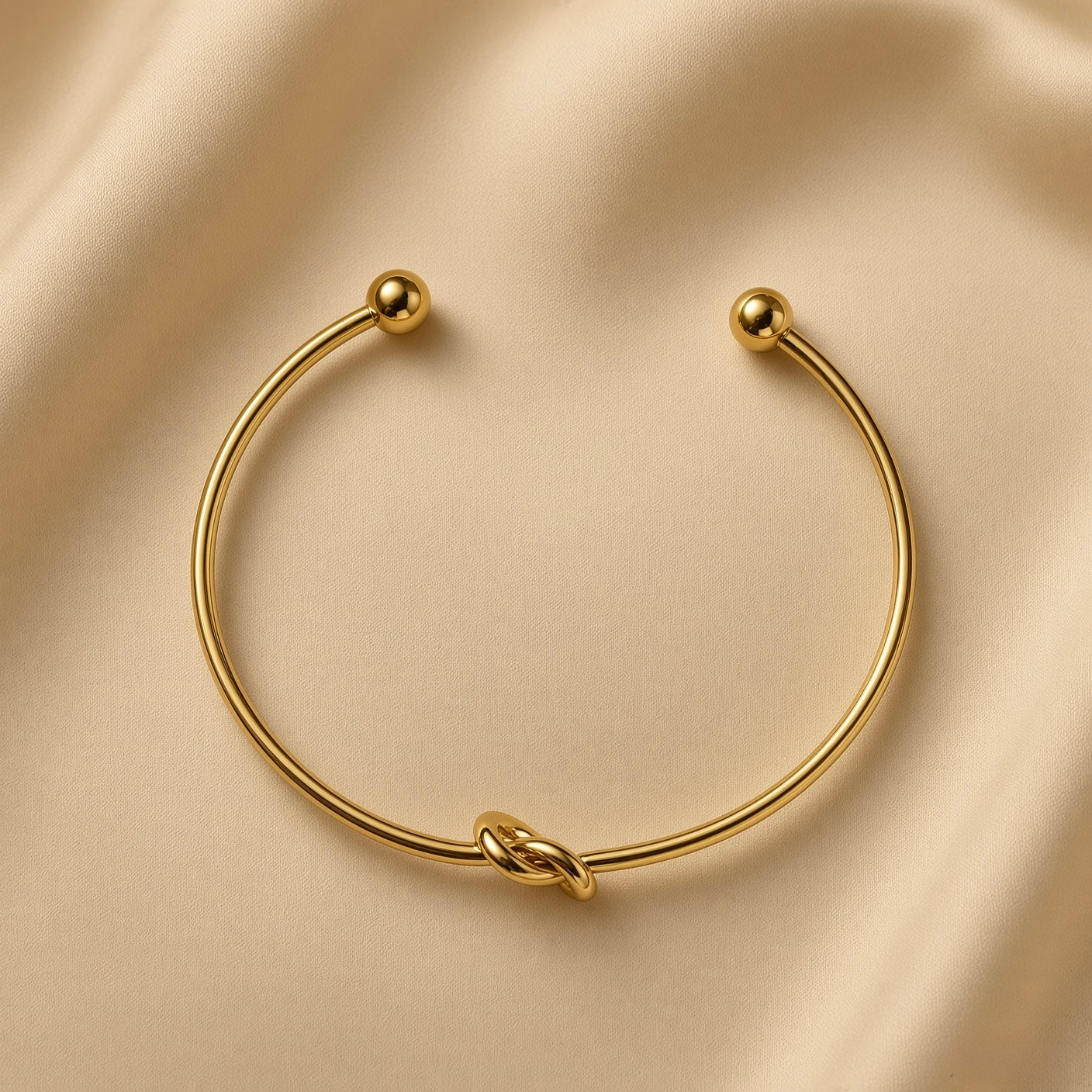 Nœud d’Amour Love Knot Ball-End Cuff Bangle - overhead view on champagne silk showing complete open cuff bangle: smooth round-wire gold arc with love knot formed in wire at base centre and two smooth polished gold ball finials at open ends