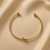 Nœud d’Amour Love Knot Ball-End Cuff Bangle - overhead view on champagne silk showing complete open cuff bangle: smooth round-wire gold arc with love knot formed in wire at base centre and two smooth polished gold ball finials at open ends