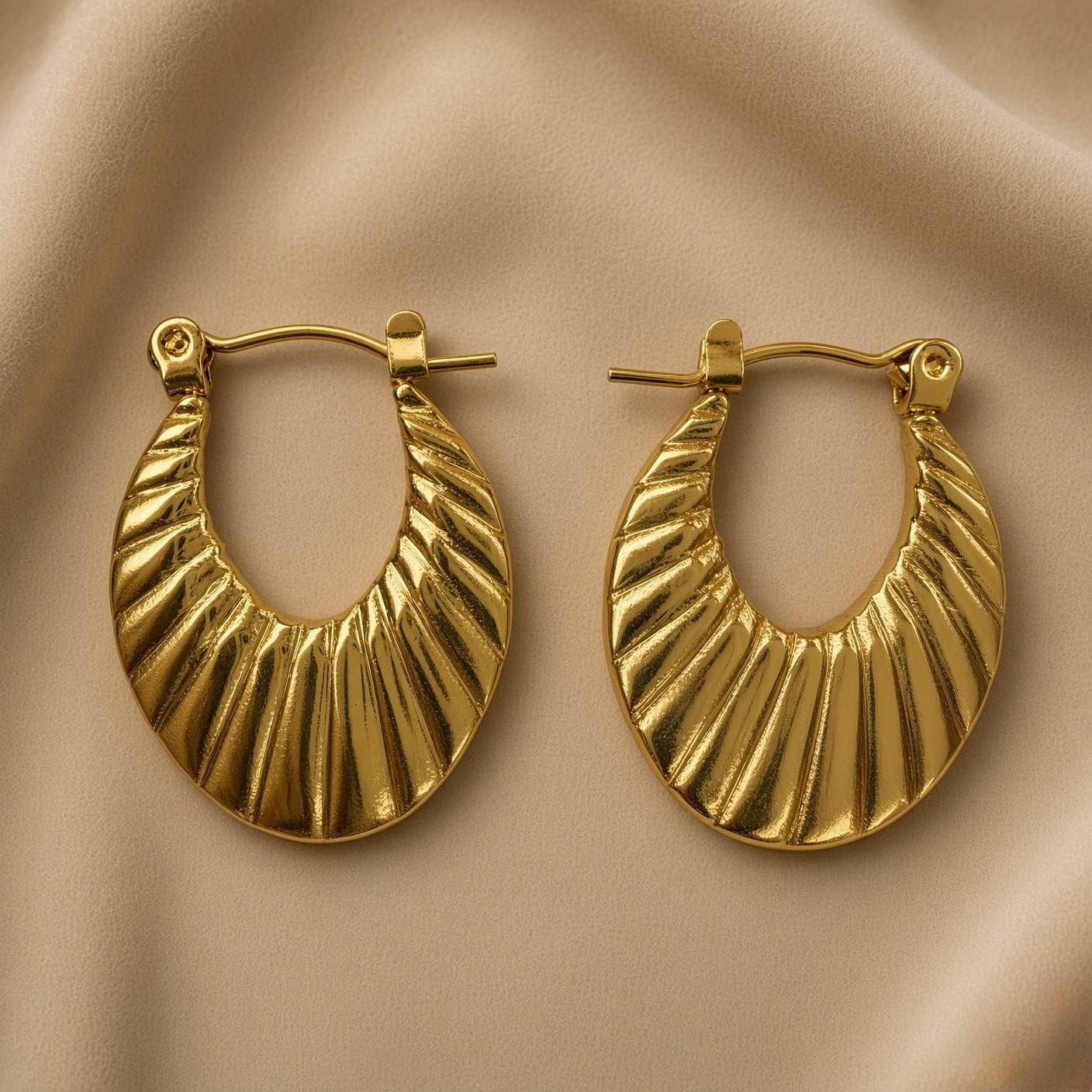 Soleil Fluted Oval Hoop Earrings - pair close-up on champagne silk showing diagonal fluted ribbing detail: deep parallel rounded ridges running upper-left to lower-right across oval hoop face with warm gold highlights on raised ridges and deep shadow grooves between, hinged latch-back clasp detail