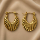 Soleil Fluted Oval Hoop Earrings - pair close-up on champagne silk showing diagonal fluted ribbing detail: deep parallel rounded ridges running upper-left to lower-right across oval hoop face with warm gold highlights on raised ridges and deep shadow grooves between, hinged latch-back clasp detail