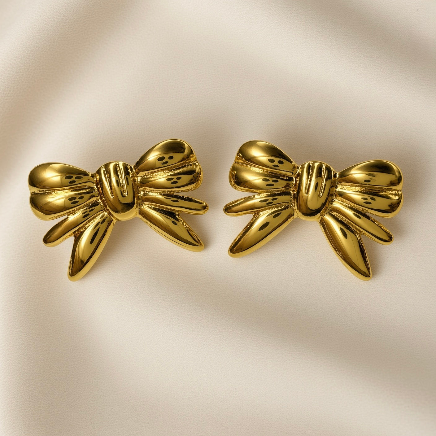 Ruban Puffy Bow Stud Earrings - pair front flat lay on ivory silk showing both large three-dimensional puffy bow stud earrings in warm 18K gold: two rounded lobes at top, tightly gathered ribbed knot at centre with three horizontal grooves, four splayed ribbon tail strands fanning outward and downward