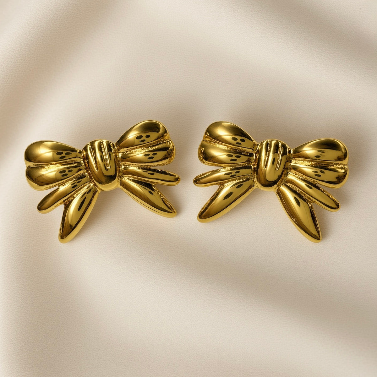Ruban Puffy Bow Stud Earrings - pair front flat lay on ivory silk showing both large three-dimensional puffy bow stud earrings in warm 18K gold: two rounded lobes at top, tightly gathered ribbed knot at centre with three horizontal grooves, four splayed ribbon tail strands fanning outward and downward