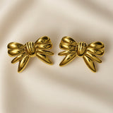 Ruban Puffy Bow Stud Earrings - pair front flat lay on ivory silk showing both large three-dimensional puffy bow stud earrings in warm 18K gold: two rounded lobes at top, tightly gathered ribbed knot at centre with three horizontal grooves, four splayed ribbon tail strands fanning outward and downward