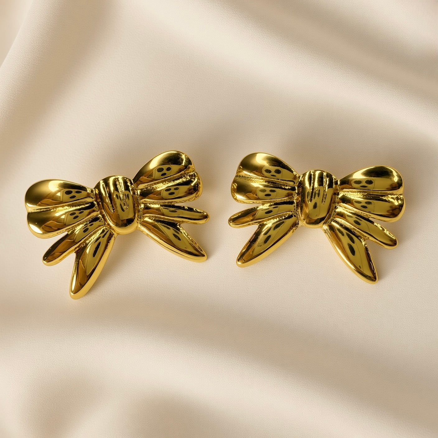 Ruban Puffy Bow Stud Earrings - pair front flat lay on ivory silk showing both large three-dimensional puffy bow stud earrings in warm 18K gold from slightly different angle: rounded lobes, ribbed central knot and four splayed ribbon tails with deep shadow recesses between elements, mirror-polish warm gold surface
