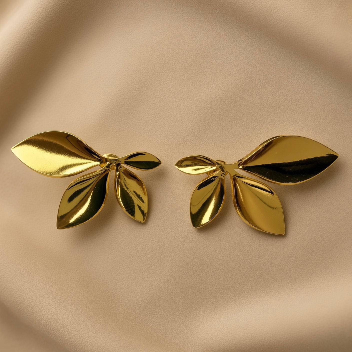 Feuilles d’Or Multi-Leaf Cluster Stud Earrings - pair front flat lay on champagne silk showing both five-leaf cluster stud earrings in warm 18K gold: one large dominant leaf extending horizontally outer, one medium leaf upward outer, one small pointed leaf top centre, two narrow leaves fanning downward, all radiating from single central point with deep shadow recesses between leaves
