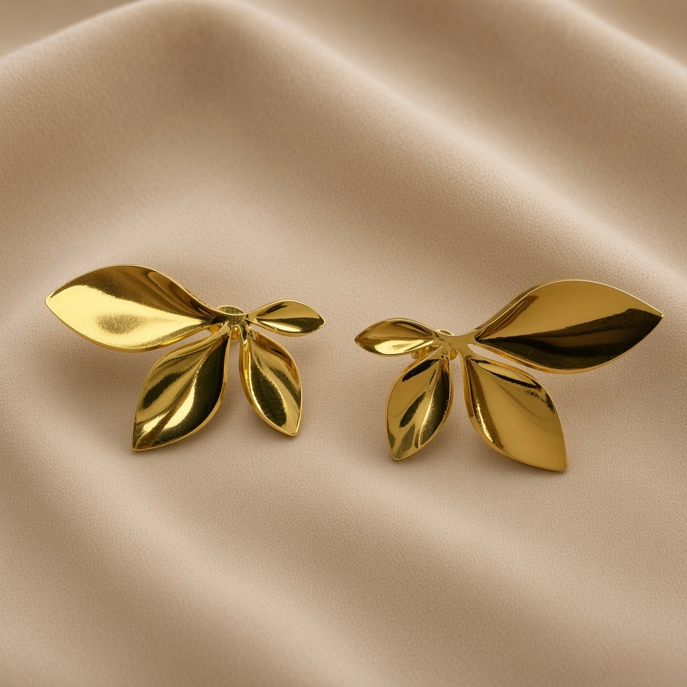 Feuilles d’Or Multi-Leaf Cluster Stud Earrings - pair three-quarter view on champagne silk showing both five-leaf cluster stud earrings in warm 18K gold from slightly elevated angle, showing three-dimensional depth of leaf forms: smooth mirror-polished convex outer surfaces, concave inner faces and deep shadow recesses between leaves at central meeting point