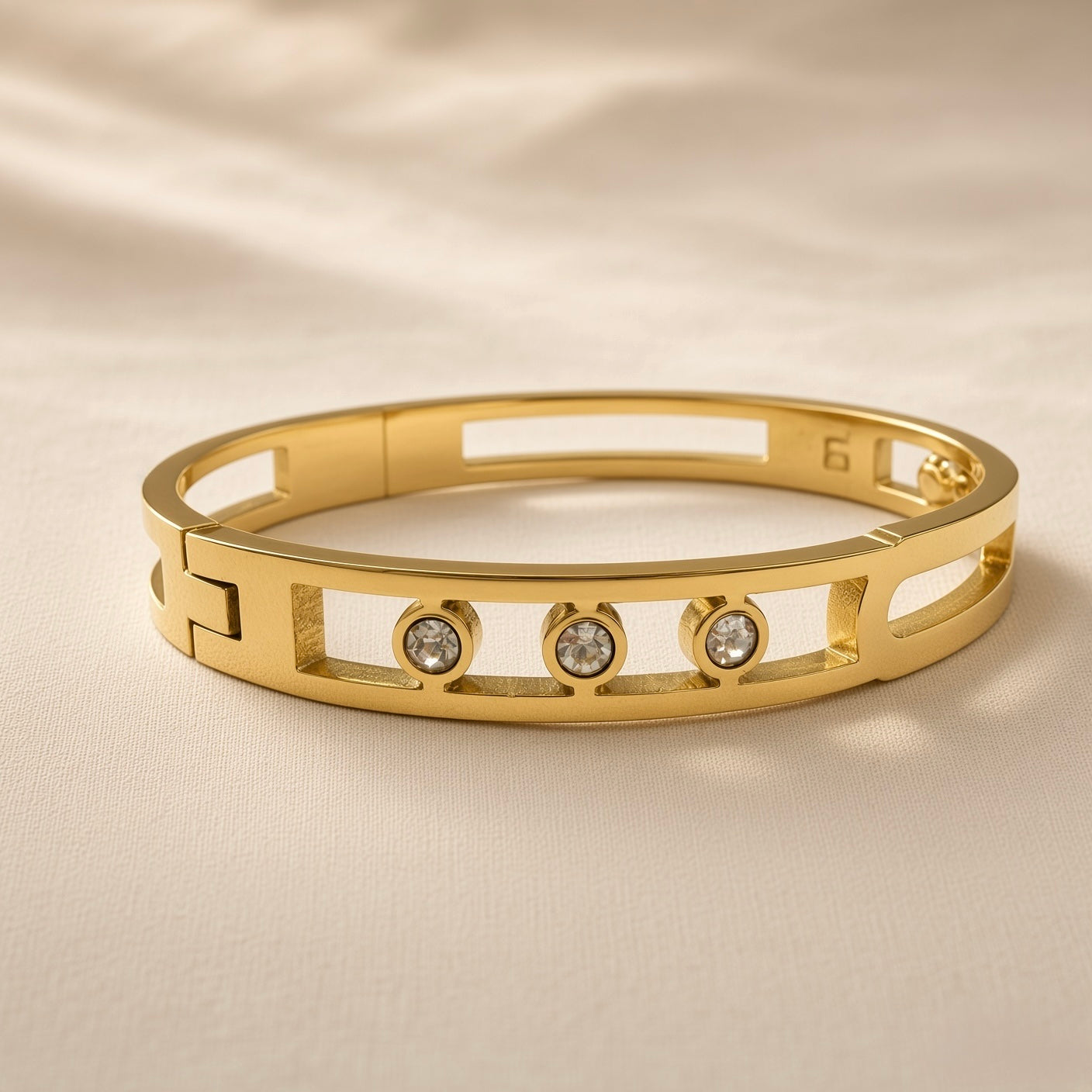 Arcade Crystal Lattice Hinged Bangle Set - crystal bangle front close-up on ivory silk showing three bezel-set round crystal zirconia stones in smooth cylindrical gold bezels within open-work geometric lattice of alternating rectangular slot and oval stadium cut-outs with solid gold bridges, hinged clasp mechanism visible at back left