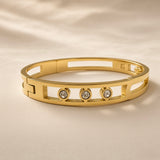Arcade Crystal Lattice Hinged Bangle Set - crystal bangle front close-up on ivory silk showing three bezel-set round crystal zirconia stones in smooth cylindrical gold bezels within open-work geometric lattice of alternating rectangular slot and oval stadium cut-outs with solid gold bridges, hinged clasp mechanism visible at back left