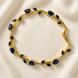 Saphir Vigne Wave Link Sapphire Crystal Bracelet - circular flat lay on ivory silk showing complete bracelet in full circle: sinuous gold wave vine links with alternating large oval deep royal sapphire blue crystals in four-prong ball-tip settings and paired marquise navette sapphire blue crystals on wave curves, box clasp with safety tab visible at right