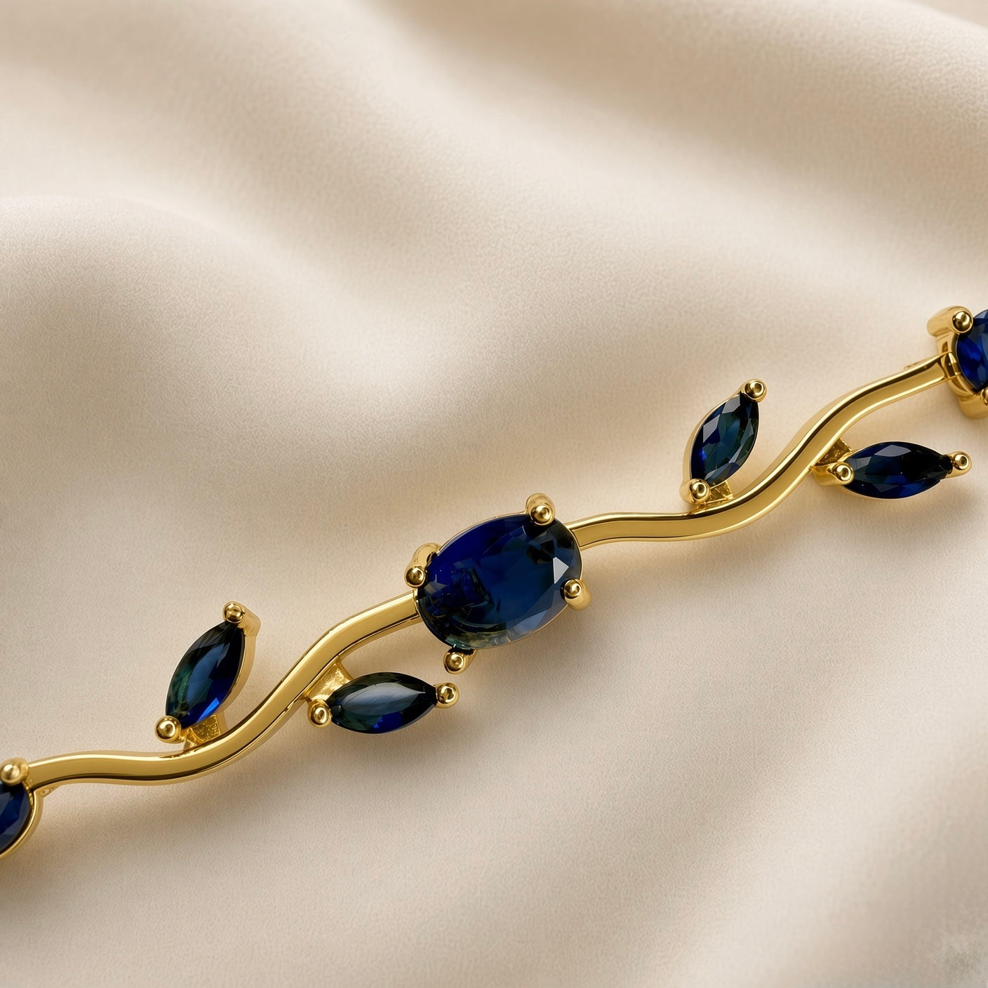 Saphir Vigne Wave Link Sapphire Crystal Bracelet - macro close-up on ivory silk showing three links in detail: large oval deep sapphire blue crystal in four-prong ball-tip gold setting at centre, paired marquise navette sapphire blue crystals in two-prong point-tip settings on sinuous S-curve gold wave links above and below, deep royal blue crystal colour and gold ball-tip prong detail clearly visible
