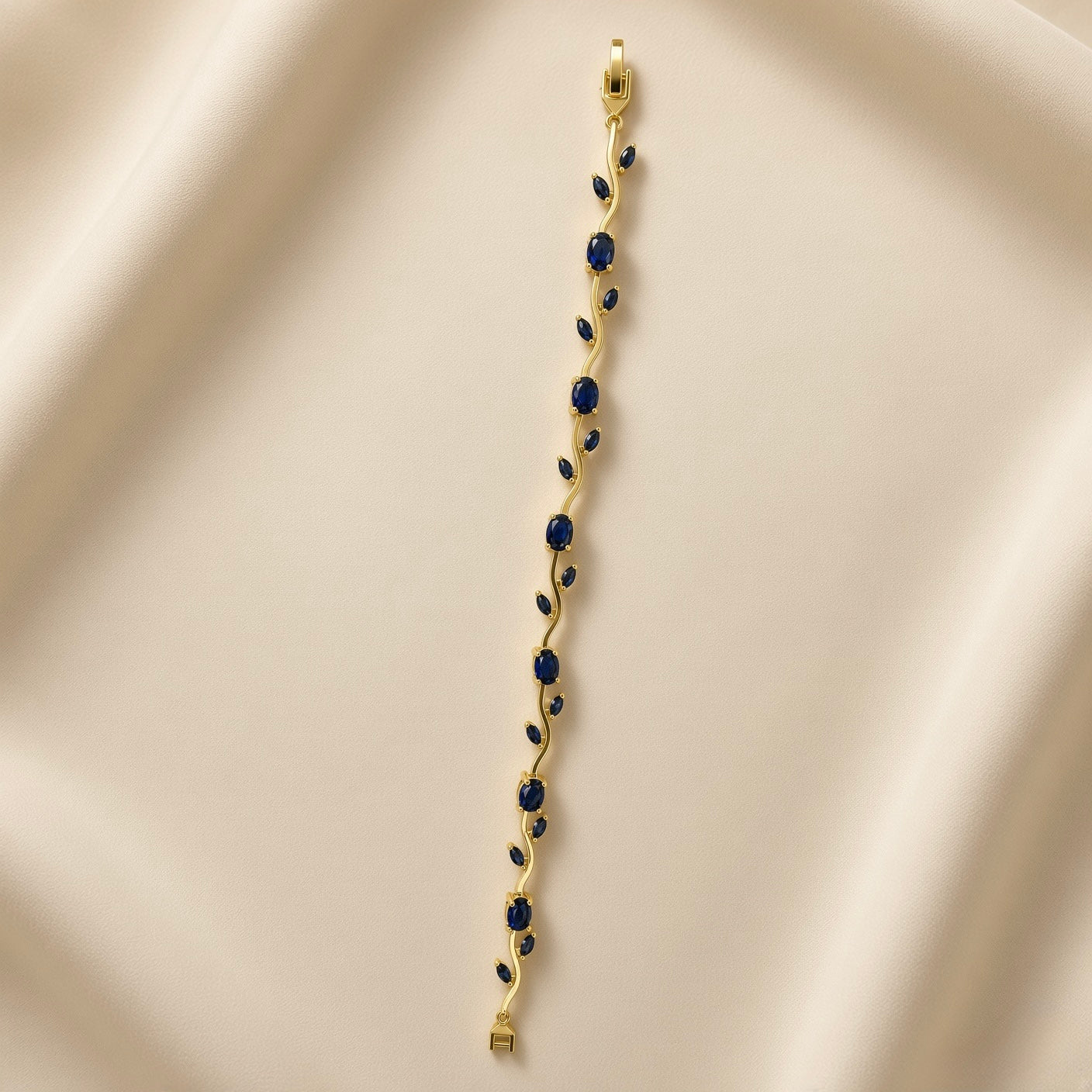 Saphir Vigne Wave Link Sapphire Crystal Bracelet - full length flat lay on ivory silk showing complete bracelet laid straight: continuous sinuous S-curve gold wave links set with alternating large oval deep sapphire blue crystals in four-prong ball-tip settings and paired marquise sapphire blue crystals in two-prong settings, box clasp with safety tab at top