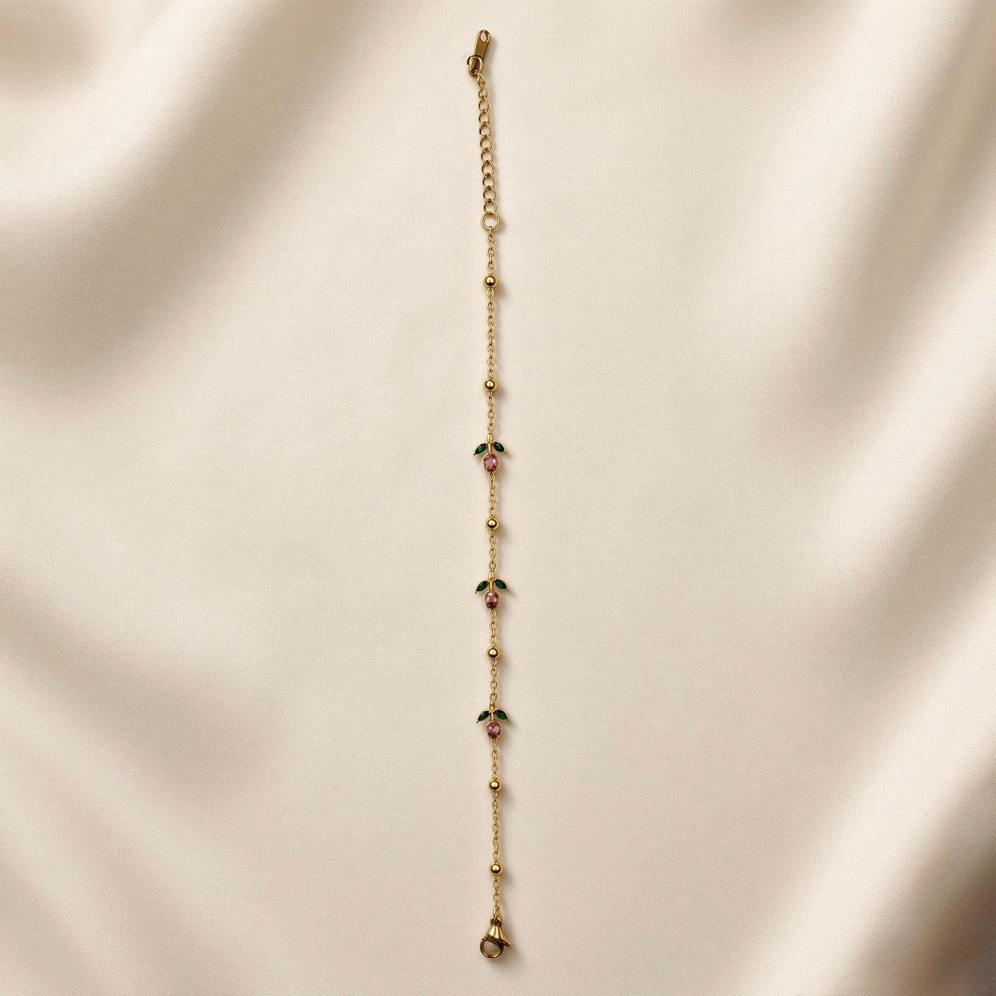 Rosette Bud Station Chain Bracelet - full length flat lay on champagne silk with natural light shadow showing complete bracelet: fine gold cable chain with gold ball stations and three rose bud charm stations with round rose-pink crystals and dark green enamel marquise sepals, lobster clasp and curb-link extender chain at top