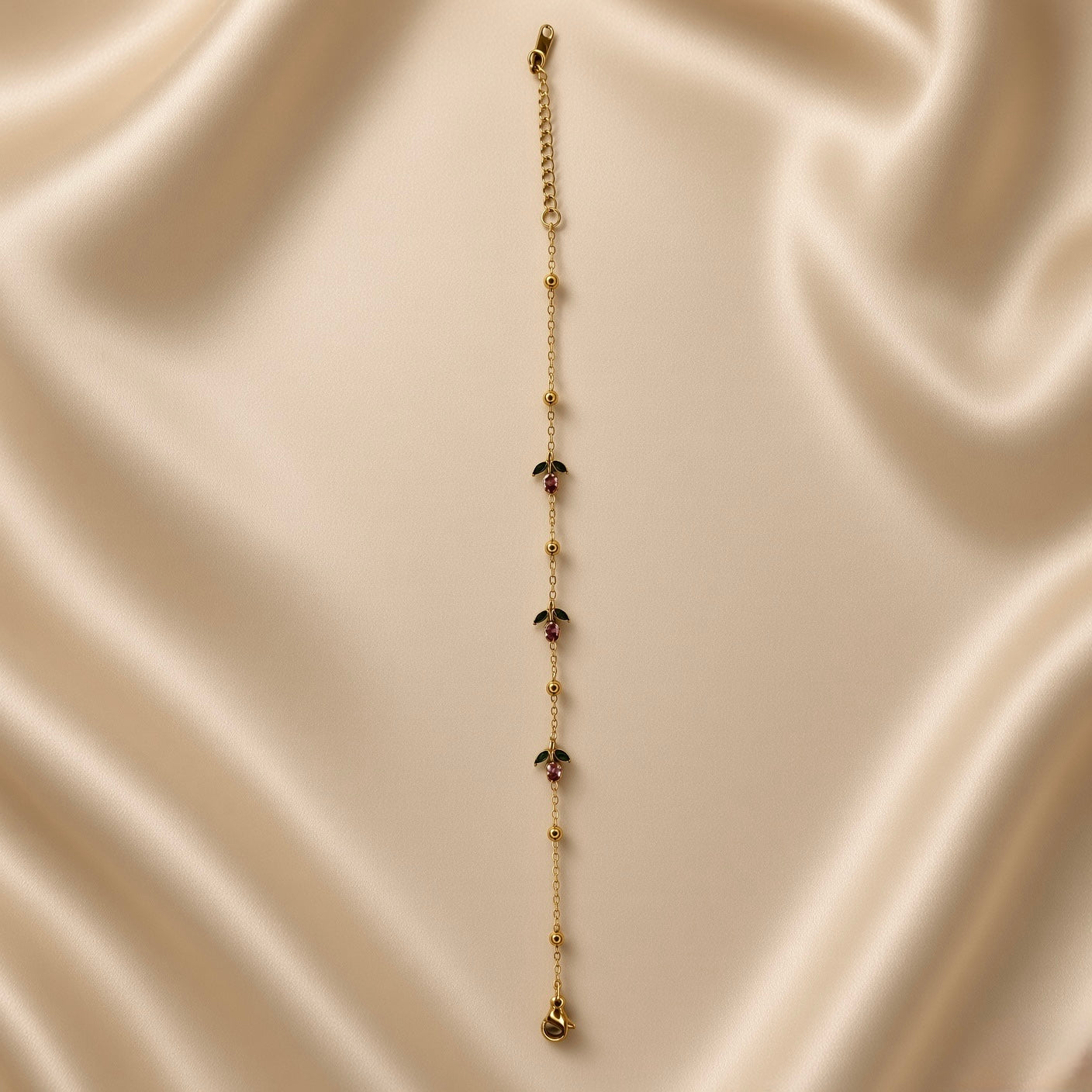 Rosette Bud Station Chain Bracelet - full length flat lay on ivory silk showing complete bracelet laid straight: fine gold oval-link cable chain with smooth gold ball stations at regular intervals and three rose bud charm stations showing round rose-pink faceted crystals with two dark green enamel marquise sepal elements, lobster clasp and curb-link extender chain at top