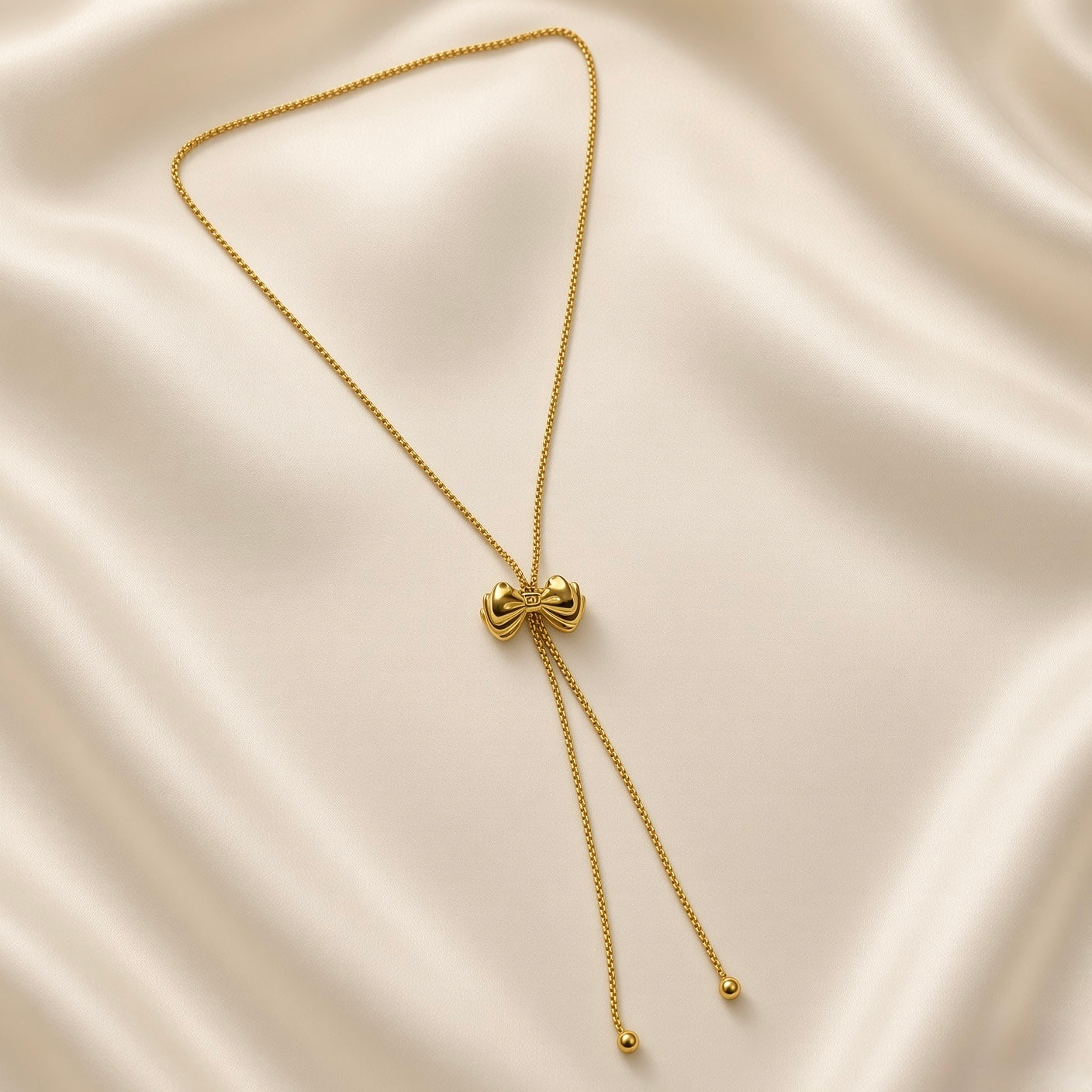 Grand Ruban Bow Lariat Box Chain Necklace - full flat lay on ivory silk showing complete necklace: continuous square-link gold box chain loop with large three-dimensional puffy bow slider at centre, two tail chains descending with smooth polished gold ball finials at ends, bow showing two rounded lobes, ribbed knot and four splayed ribbon tail strands