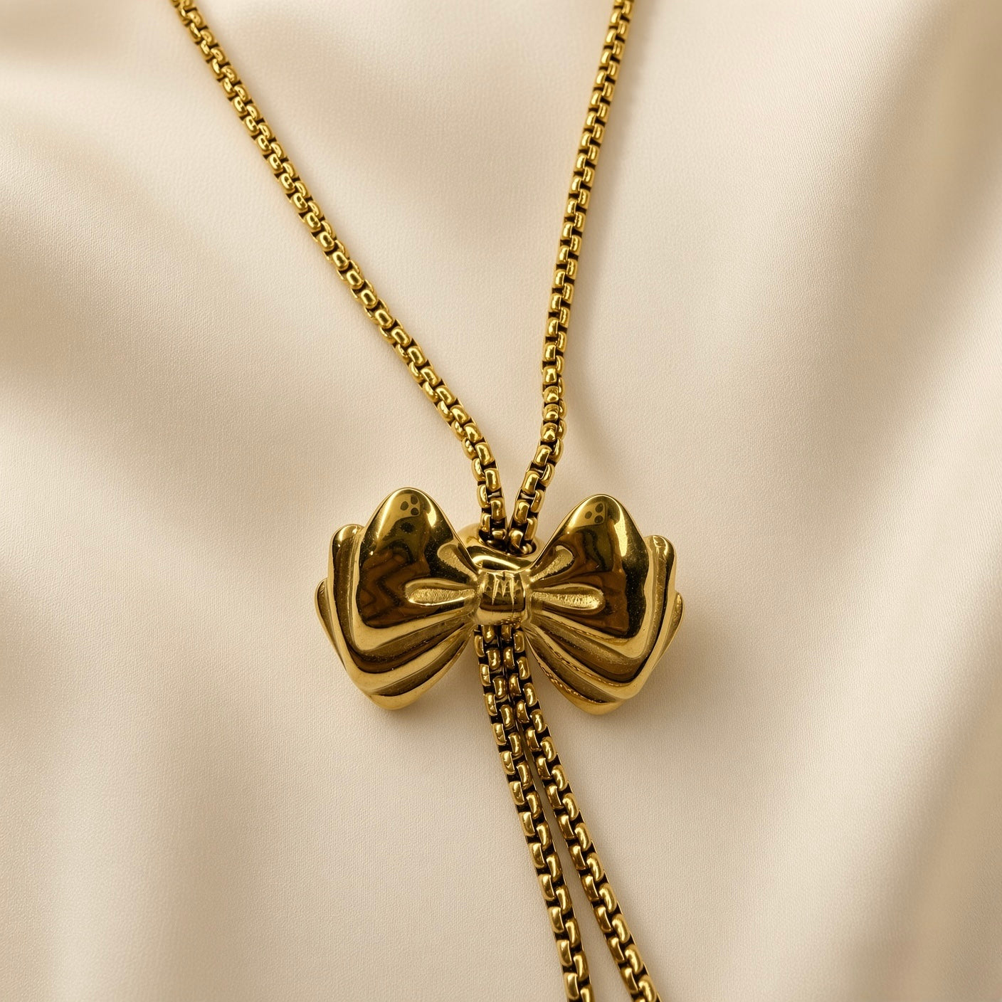 Grand Ruban Bow Lariat Box Chain Necklace - macro close-up on champagne silk showing puffy bow slider in extraordinary detail: two large rounded mirror-polished gold lobes, tightly gathered ribbed central knot with multiple deep horizontal grooves, four splayed ribbon tail strands fanning outward, square-link box chain threaded through bow knot channel, two tail chains descending below