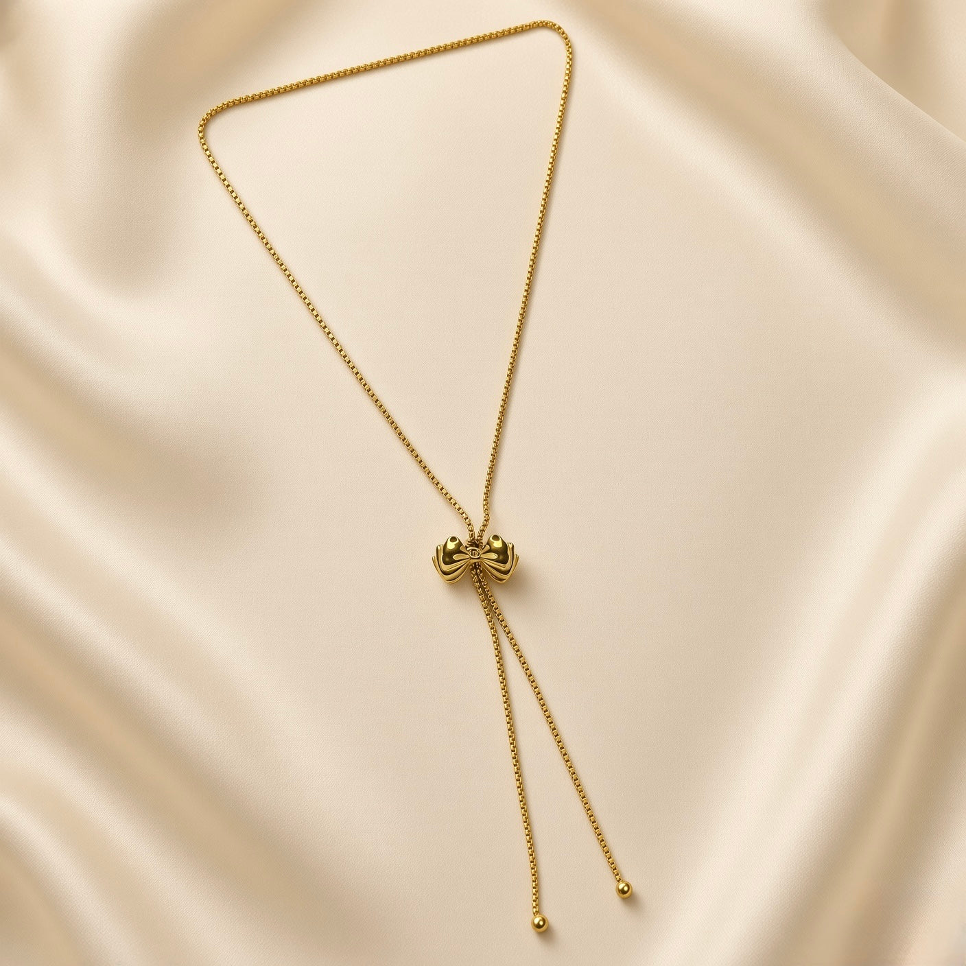 Grand Ruban Bow Lariat Box Chain Necklace - full flat lay on ivory silk showing complete necklace from slightly different angle: continuous square-link gold box chain loop with large puffy bow slider at centre showing two rounded lobes and ribbed knot, two parallel tail chains with smooth polished gold ball finials descending below bow