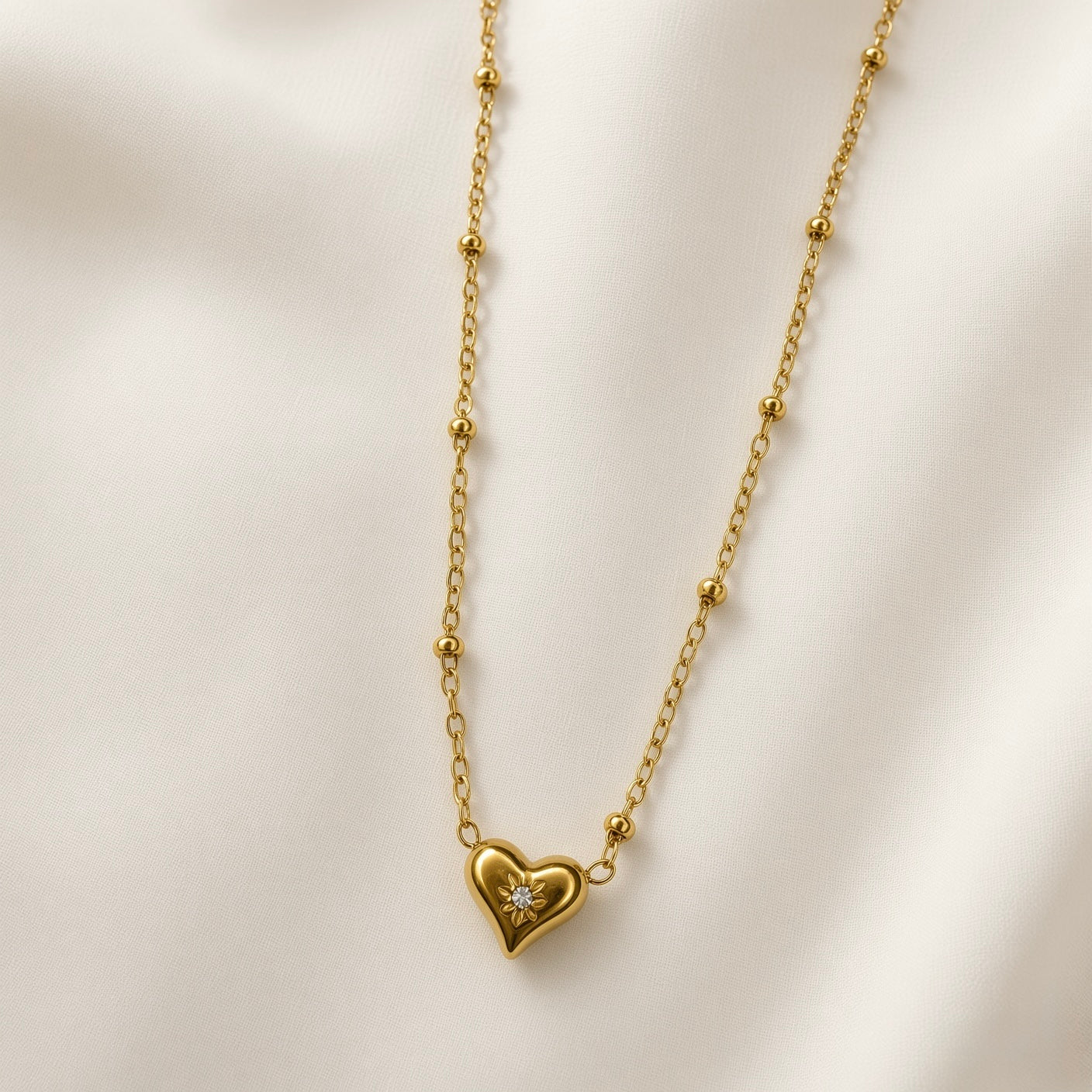 Étoile du Cœur Puffy Heart Satellite Chain Necklace - close-up flat lay on ivory silk showing three-dimensional puffy heart pendant in warm 18K gold with single round brilliant crystal zirconia in eight-point star sunburst bezel setting at centre of heart face, fine satellite chain with smooth gold ball stations visible on both sides