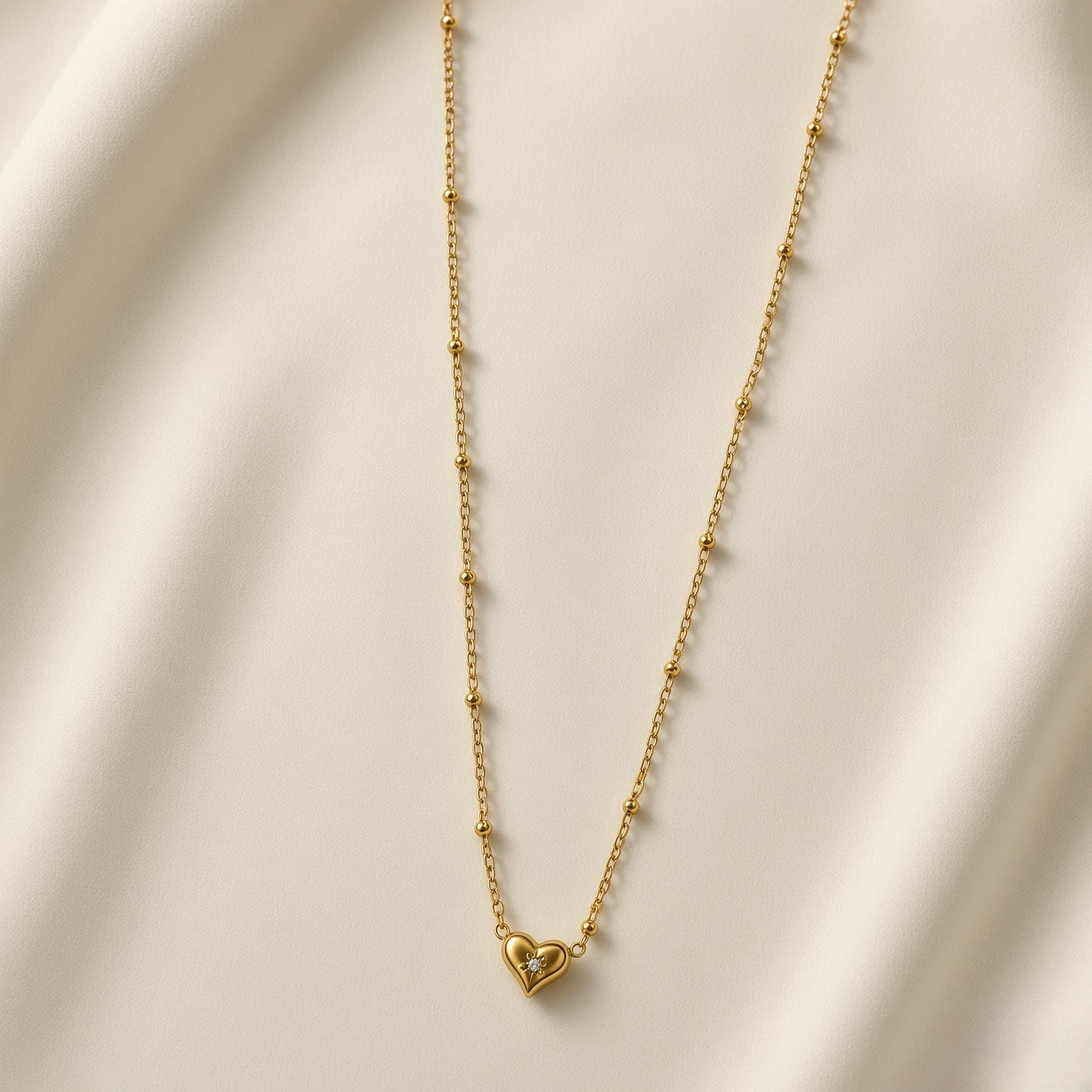 Étoile du Cœur Puffy Heart Satellite Chain Necklace - full flat lay on ivory silk showing complete necklace: fine satellite chain with smooth gold ball stations at regular intervals, three-dimensional puffy gold heart pendant with star crystal centre at bottom of chain loop