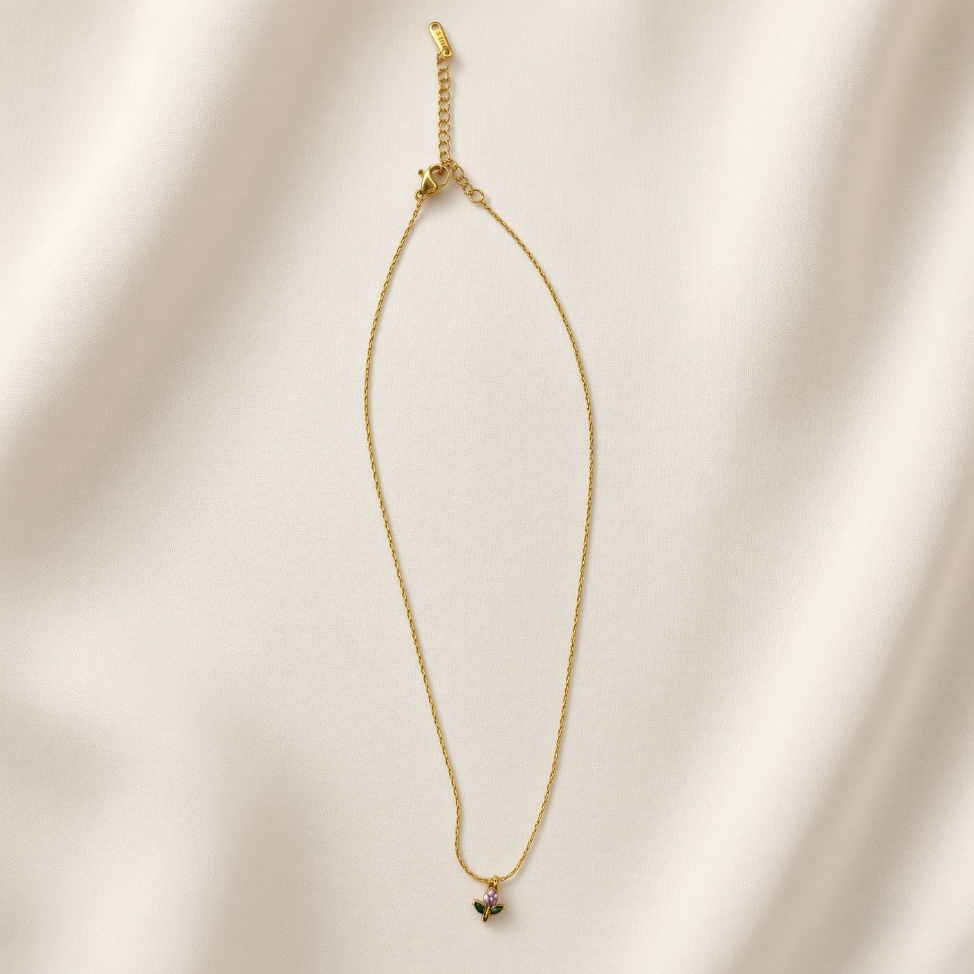 Tulipe Crystal Bud Pendant Necklace - full length flat lay on ivory silk showing complete necklace laid straight: fine twisted oval-link gold cable chain with tulip bud pendant at base showing pear pink-lilac crystal bud and two marquise deep green crystal leaves on gold stem, lobster clasp and extender chain with small branded tag at top