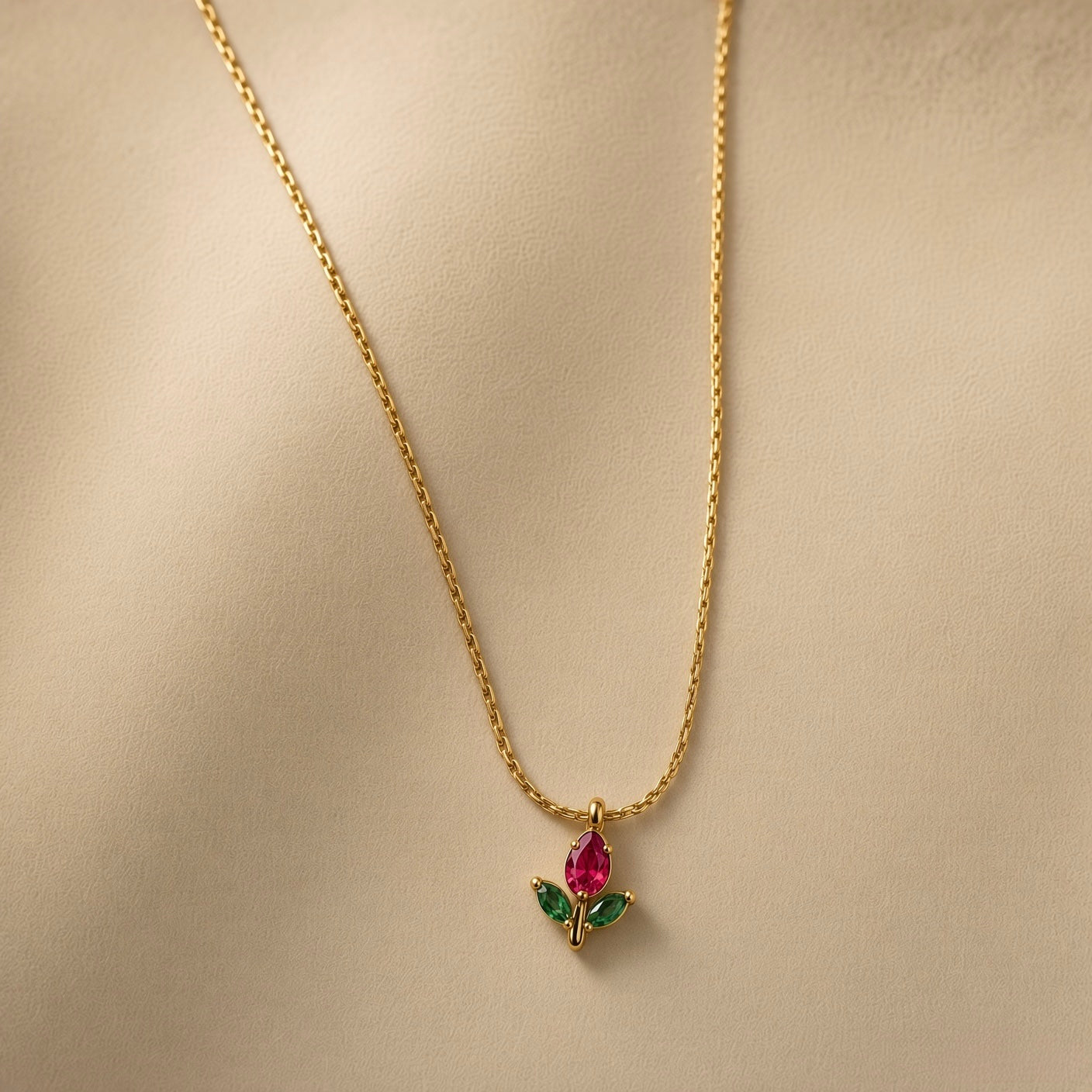 Tulipe Rouge Crystal Bud Pendant Necklace - three-quarter close-up on warm beige background showing red tulip bud pendant in full detail: pear-cut deep ruby red crystal bud in minimal gold bezel set point-downward, two marquise-cut deep forest green crystal leaves set horizontally outward from fine gold stem, smooth gold bail at top, fine twisted gold cable chain above
