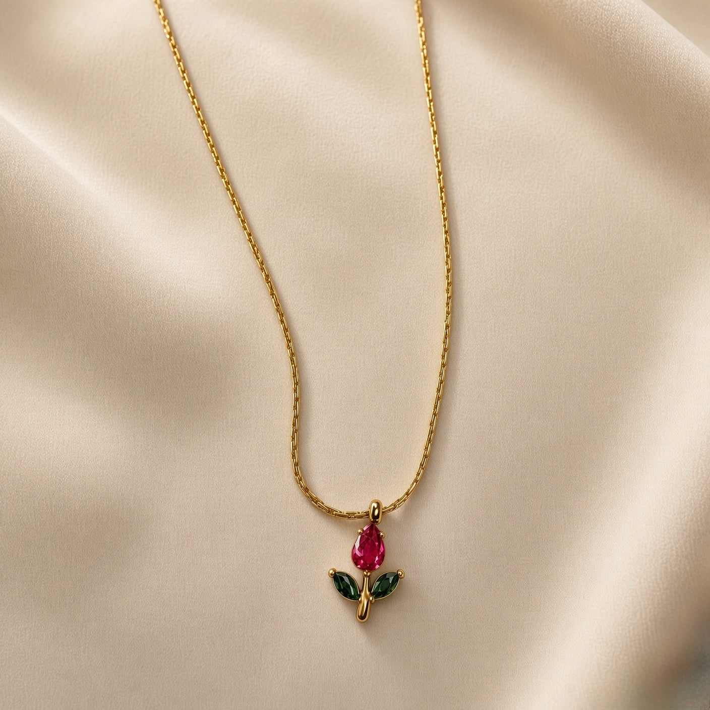 Tulipe Rouge Crystal Bud Pendant Necklace - close-up on champagne silk showing red tulip bud pendant detail: pear-cut deep ruby red crystal bud above two marquise-cut deep forest green crystal leaves on fine gold stem, fine twisted cable chain visible above smooth gold bail, deep red crystal colour and green leaf contrast clearly visible