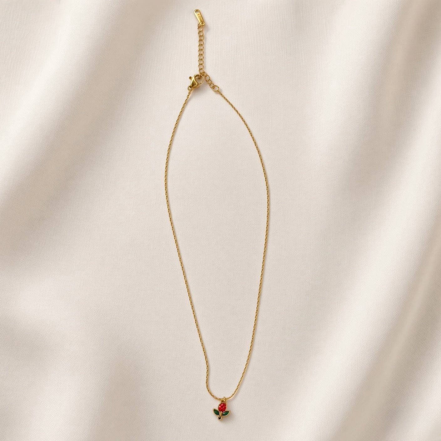 Tulipe Rouge Crystal Bud Pendant Necklace - full length flat lay on ivory silk showing complete necklace laid straight: fine twisted oval-link gold cable chain with red tulip bud pendant at base showing pear-cut deep ruby red crystal bud and two marquise deep forest green crystal leaves on gold stem, lobster clasp and extender chain with small branded tag at top