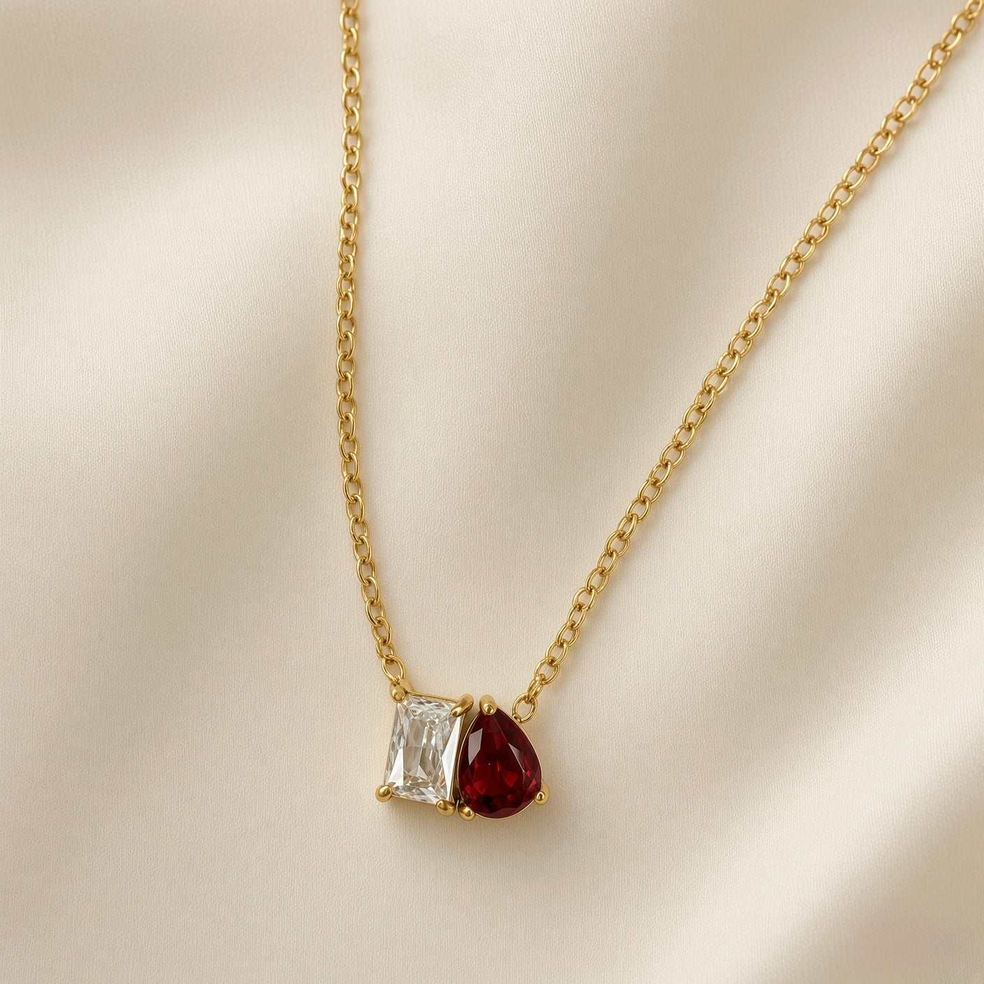 Toi et Moi Diamond Ruby Two-Stone Pendant Necklace - close-up on champagne silk showing toi et moi pendant in full detail: princess-cut diamond-clear white crystal zirconia in four-prong gold setting (left) touching pear-cut deep ruby red crystal zirconia in three-prong gold setting (right), two stones sharing edge in single pendant, fine oval-link gold cable chain above