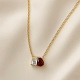 Toi et Moi Diamond Ruby Two-Stone Pendant Necklace - close-up on champagne silk showing toi et moi pendant in full detail: princess-cut diamond-clear white crystal zirconia in four-prong gold setting (left) touching pear-cut deep ruby red crystal zirconia in three-prong gold setting (right), two stones sharing edge in single pendant, fine oval-link gold cable chain above