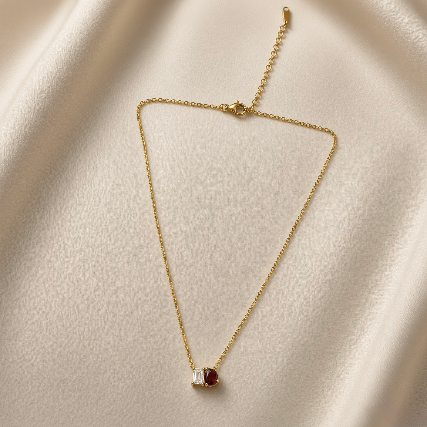 Toi et Moi Diamond Ruby Two-Stone Pendant Necklace - full length flat lay on champagne silk showing complete necklace from slightly different angle: fine oval-link gold cable chain with toi et moi pendant showing princess-cut white crystal zirconia and pear-cut deep ruby red crystal zirconia in shared gold setting, lobster clasp and curb-link extender chain at top