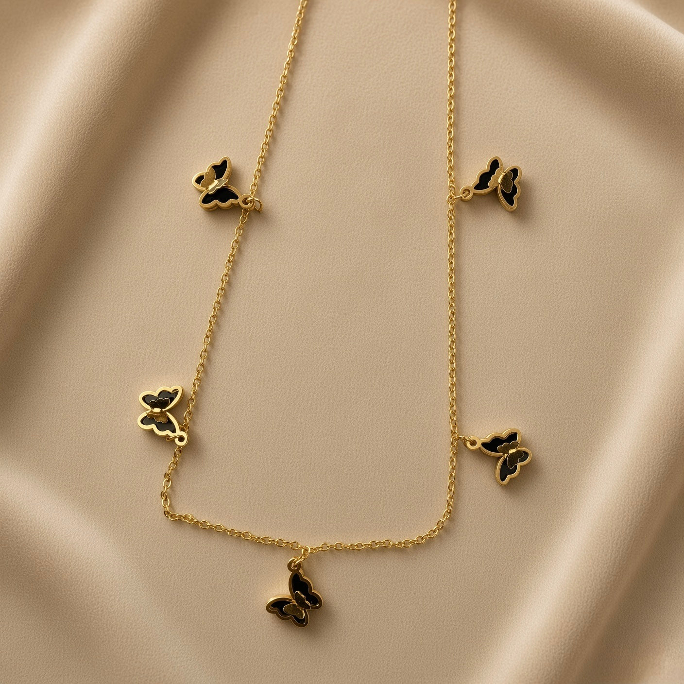 Papillon Noir Black Enamel Butterfly Charm Necklace - flat lay on champagne silk showing complete necklace in V silhouette: fine gold oval-link cable chain with five graduated black enamel butterfly charms at asymmetric intervals, two small butterflies upper sides, two medium butterflies lower sides, one large butterfly central drop pendant at bottom, deep black enamel fill with warm gold border outline on each butterfly